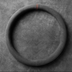 May include: A circular steering wheel cover made of dark gray suede-like material. The cover features a smooth, padded rim with a single vertical red stitched accent mark positioned at the top center of the circular frame.