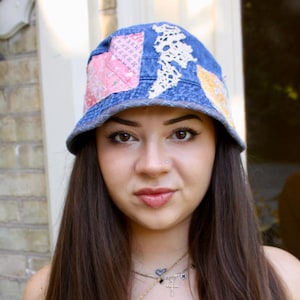 以下が含まれることがあります： ピンク、白、黄色のパッチワークが施された青いデニムのバケットハット。 レースパネルが付いています。 長い茶色の髪の人がかぶっています。