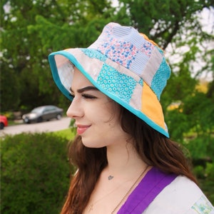 Op de afbeelding: Een vrouw draagt ​​een patchwork bucket hat met een blauwe rand en een kroon met een geel, wit en roze patroon. Ze draagt ​​een wit en paars jack met een bloemenpatroon.