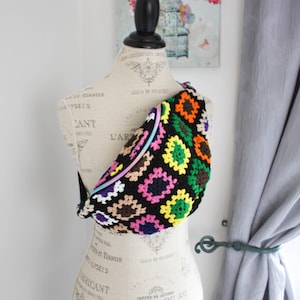 May include: A colorful crocheted fanny pack with a rainbow zipper. The fanny pack is on a white mannequin with a floral print background.