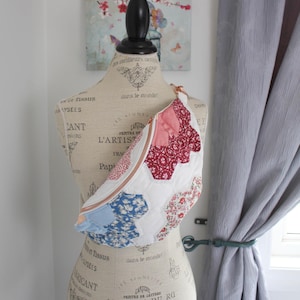 May include: A crossbody bag featuring a patchwork design of white, red, and blue floral fabric hexagons. The bag has a gold-toned zipper closure and is displayed on a mannequin torso printed with French text and decorative script patterns.