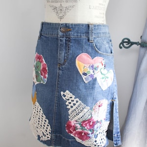 May include: A denim skirt with colourful fabric patches and lace accents. The patches include floral and heart shapes. The skirt has a slit on the side and is displayed on a mannequin.