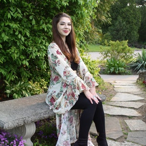 Könnte beinhalten: Ein Kimono mit Blumenmuster und Glockenärmeln, mit cremefarbenem Hintergrund und einem Muster aus roten, rosa und grünen Blumen. Der Kimono wird über einem schwarzen Oberteil und schwarzen Leggings getragen. Die Person trägt schwarze High Heels mit offenem Zehenbereich.