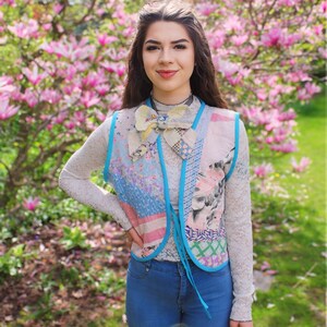 以下が含まれることがあります： 青いデニムのベストに、花柄のパッチワークのデザインと青い蝶ネクタイをつけた女性。ピンクの花が咲いた木の前に立っている。