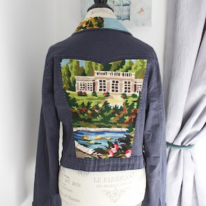 May include: Dark blue denim jacket featuring a detailed needlepoint design on the back. The needlepoint shows a landscape scene with a building, trees, and water. The jacket has a classic collar and button fastenings.