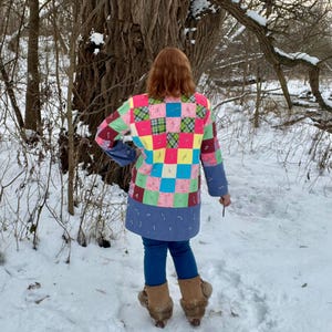May include: A vibrant patchwork jacket featuring a mix of square patterns in pink, green, blue, and yellow. The jacket has blue sleeves and a blue hemline. The person is wearing blue jeans and brown boots in a snowy outdoor setting.