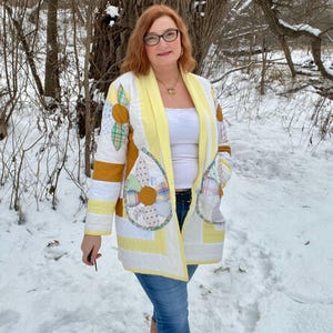 May include: A patchwork jacket in shades of yellow, white, and mustard, with floral and geometric patterns. The open-front jacket has large pockets and is worn over a white top and blue jeans. The background is a snowy outdoor scene.