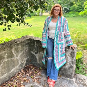 May include: A long, open-front crochet cardigan in a granny square pattern. The colors include turquoise, pink, white, and purple. The cardigan is worn over a white top and blue jeans. The background features a stone wall and green foliage.