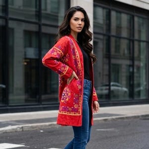 May include: A long-sleeved red jacket featuring intricate purple, orange, and yellow embroidery patterns, worn over a black top and blue denim flare jeans with black leather boots, creating a layered outfit with visible front pockets.