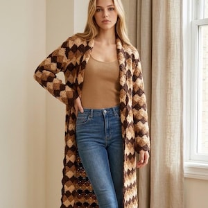 May include: A long, open-front crochet cardigan featuring a repeating diamond pattern in shades of dark brown, medium brown, and cream. The garment is worn over a tan tank top, blue denim jeans, and brown suede ankle boots with a block heel.