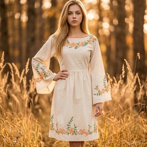 May include: A cream-coloured dress featuring long, flared sleeves and a round neckline. The garment is decorated with embroidered floral patterns in orange and green thread, which are positioned along the neckline, the sleeves, and the hemline.