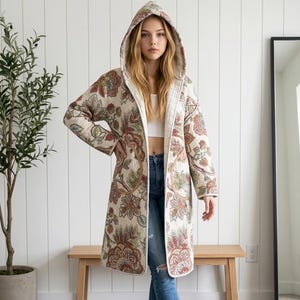 May include: A long-sleeved, knee-length hooded coat featuring a cream-coloured base with a multi-coloured floral pattern in shades of red, green, and brown. The coat has a quilted texture, an open front, and two large circular patch pockets on the front.
