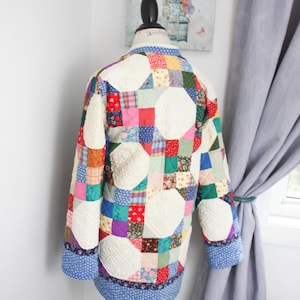 May include: A long-sleeved jacket constructed from a patchwork quilt featuring a variety of colorful fabric squares in red, blue, green, yellow, and white. The garment includes large cream-colored circular quilted panels and blue patterned cuffs.