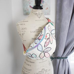 May include: A white fabric crossbody bag featuring a colorful leaf pattern with black stitched outlines. The bag has a bright blue zipper closure and a coral-colored strap. It is displayed on a beige dress form with French typography print.