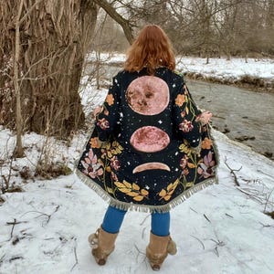 Pode incluir: Um quimono preto com um design celestial com luas rosa e motivos florais. O casaco, com bainha com franjas, é usado sobre leggings azuis e botas castanhas. O fundo mostra uma paisagem nevada com um rio e árvores.