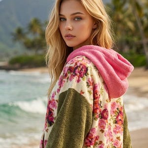 Könnte beinhalten: Ein Fleece-Kapuzenpullover mit cremefarbener Basis und einem lebhaften rosa-roten Blumenmuster, akzentuiert durch einfarbige olivgrüne Ärmel und eine texturierte, leuchtend rosa Kapuze. Das Kleidungsstück besteht aus weichem, flauschigem Material.