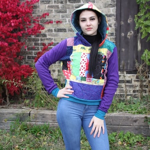 Puede incluir: Una mujer con una sudadera con capucha de patchwork de colores con una capucha azul turquesa. La sudadera tiene un diseño acolchado y está hecha de diferentes telas. La mujer lleva pantalones vaqueros azules.