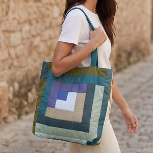 May include: A quilted tote bag featuring a geometric log cabin pattern in shades of olive green, teal, lavender, light blue, and beige. The bag has two matching fabric shoulder straps and a rectangular shape with visible stitching throughout the design.