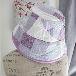 Peut inclure: Un chapeau seau en patchwork violet et blanc avec un motif à carreaux bleu clair et blanc. Le chapeau est en tissu et a une large bordure.