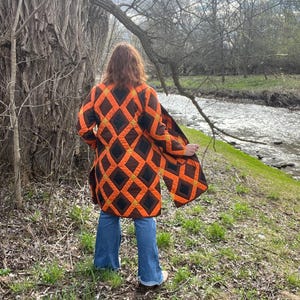 May include: A knee-length quilted coat in orange and black, featuring a diamond pattern. The coat is open, with long sleeves. The person is wearing blue jeans and white shoes. The background shows a river and trees.
