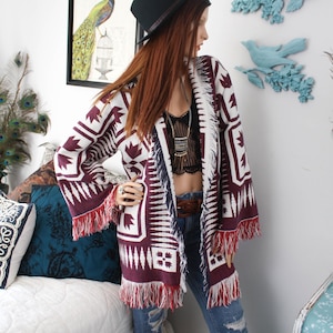 May include: A long-sleeved open-front cardigan featuring a deep burgundy and white geometric pattern with fringe trim along the hem and sleeve cuffs. The garment is worn over a black lace crop top, a brown leather belt, and distressed blue jeans.