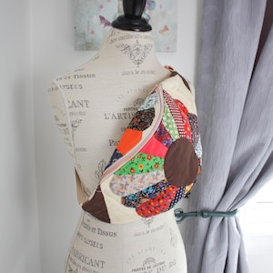 May include: A crossbody bag featuring a patchwork design of various floral and geometric fabric patterns in red, orange, green, and blue, centred with a solid brown circle. The bag has a cream-coloured strap and a zip closure across the top.