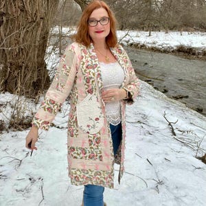 May include: A long, patchwork coat in shades of pink, cream, and green, with floral patterns. The coat has a large pocket with floral embroidery. The person is wearing blue jeans and tan boots. The background shows a snowy landscape.