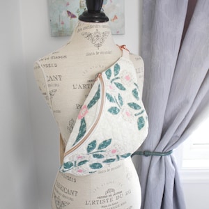 May include: A cream-coloured quilted fanny pack with a floral pattern of green leaves and pink flowers. The bag has a gold zip and a cream-coloured strap. The bag is displayed on a mannequin.