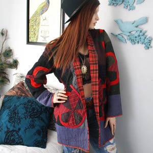 May include: A long-sleeved patchwork jacket featuring a mix of black, red, and blue fabric panels with a red plaid lining. The jacket includes circular quilted patterns, a straight hem, and an open front design worn over a black top and blue jeans.
