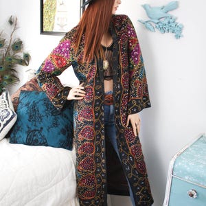 May include: A long, black embroidered duster coat with colorful paisley patterns and gold accents. The coat is worn with blue jeans and brown cowboy boots. A black hat is worn on the head. The background includes a peacock painting and blue bird wall decor.
