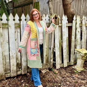 Può includere: Un lungo cappotto patchwork dai colori pastello, tra cui rosa, giallo e verde, con un design trapuntato. Il cappotto ha tasche e un bordo a righe. La persona indossa jeans blu e un top bianco. Lo sfondo include una staccionata bianca e una casetta per uccelli.