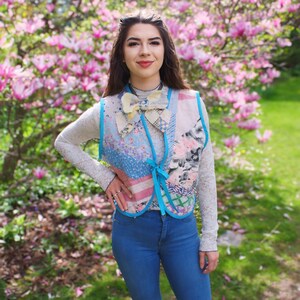 以下が含まれることがあります： ピンクの花が咲いた木の前に立っている女性。彼女は、パッチワークのデザインと青い蝶ネクタイが付いた青いデニムのベストを着ています。