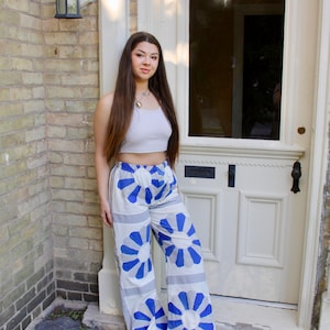 May include: White wide-leg trousers with a blue floral pattern. The design features a high waist and a striking pattern. The model is wearing a light grey crop top and black shoes. The trousers are a statement piece, perfect for a summer look.