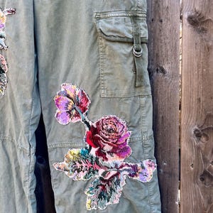 May include: Green cargo trousers with a floral embroidered patch on the right leg. The patch features a large pink rose with green leaves and smaller pink flowers.