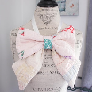 Peut inclure: Un nœud papillon fait à la main en patchwork rose, rouge et bleu, présenté sur un mannequin. Le nœud papillon est fabriqué à partir de tissu matelassé, mettant en valeur un design unique et fait main. Un accessoire décoratif.