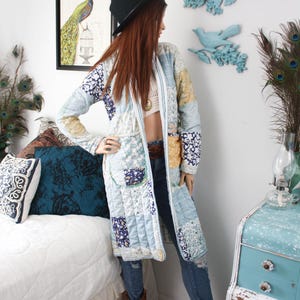 May include: A long-sleeved, knee-length quilted coat featuring a patchwork design with various light blue, white, and yellow floral and geometric patterns. The coat has a straight open front, a collarless neckline, and two large patch pockets on the front.