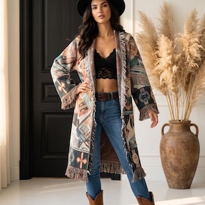 May include: A long, open-front cardigan featuring a multi-coloured woven tapestry pattern with geometric shapes and fringe trim along the edges. The garment is worn over a black lace bralette, blue denim jeans with a brown belt, and brown leather boots.