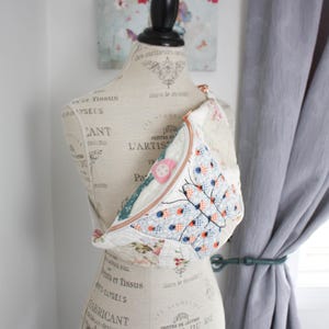 May include: A quilted crossbody bag featuring a patchwork design with floral patterns and a prominent butterfly motif in shades of blue, orange, and white. The bag has a copper-colored zipper closure and is displayed on a dress form mannequin.