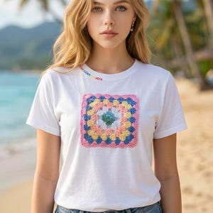 May include: A white short-sleeved t-shirt featuring a square, multi-coloured crochet granny square patch on the chest and decorative multi-coloured cross-stitching along the neckline, paired with blue denim jeans with distressed details.