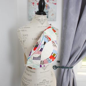 May include: A crescent-shaped crossbody bag featuring a patchwork design with various colorful fabric patterns, including floral, plaid, and animal prints, joined by white quilted stitching and secured with a light blue zipper closure.