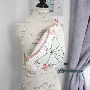 May include: A quilted crossbody bag featuring a floral pattern on a cream background with a large, embroidered light green butterfly motif on the front. The bag has a zip closure and is displayed on a dress form mannequin with printed text.