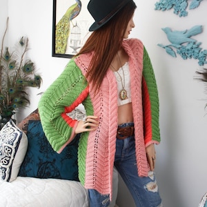 May include: A long-sleeved, open-front knit cardigan featuring vertical panels of light pink and green yarn with a textured chevron stitch pattern and thin orange accents along the sleeves, worn over a white top and distressed blue denim jeans.