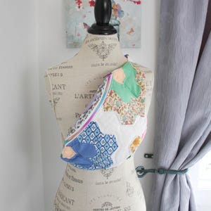 May include: A crossbody bag constructed from a patchwork of various fabric hexagons in shades of green, blue, white, and floral patterns. The bag features a prominent zipper closure along the top edge and is displayed on a dress form mannequin.