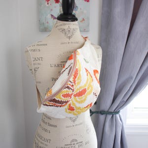 May include: A white fanny pack with a colourful floral design in orange, yellow, and brown. The bag has a light brown zip and strap. The bag is displayed on a mannequin.