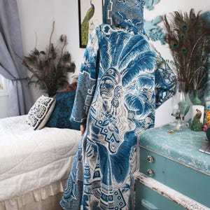 Peut inclure: Un manteau à capuche à manches longues avec un motif de tapisserie tissé dans des tons de bleu sarcelle, bleu et blanc. Le design présente un grand graphique détaillé d'une silhouette portant une coiffe à plumes élaborée et des motifs géométriques.