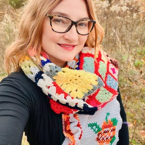 Puede incluir: Una mujer con una camisa negra y una bufanda de ganchillo de colores con un diseño de calabaza. La bufanda está hecha de cuadrados de diferentes colores y tiene un borde rizado.