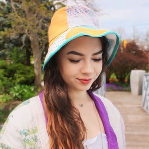 May include: A woman wearing a yellow, blue and white patterned bucket hat with a turquoise brim. She is wearing a white and purple patchwork jacket and a silver necklace.