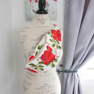 May include: White beaded fanny pack with a red floral design. The bag is shaped to fit the body and has a zipper closure. The strap is a light brown color. The bag is displayed on a mannequin.