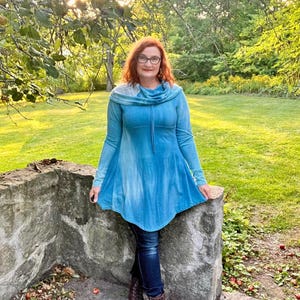 May include: A long-sleeved, blue tunic with a cowl neck and a flared hem. The tunic is paired with dark blue jeans and brown lace-up boots. The background features a stone wall, green grass, and trees.