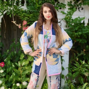 Peut inclure: Une femme portant une veste en patchwork avec des bordures roses et un pantalon blanc à motif floral bleu et rose. Elle se tient devant une clôture blanche avec de la végétation verte.